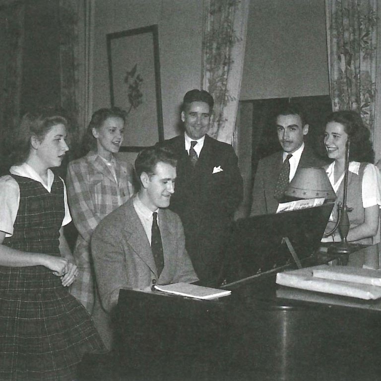 A historical photo of men and women gathered around a man playing the piano