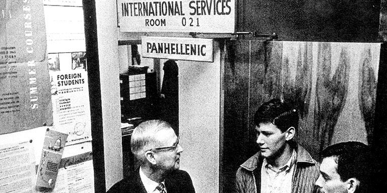 The dean and two students stand beneath a sign that reads, International Services Room 021.